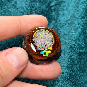 May include: A round, wooden pendant with a colorful, iridescent skull design. The skull is filled with a mosaic of vibrant colors, and the wood has a rich, brown tone. The pendant is held in a hand.