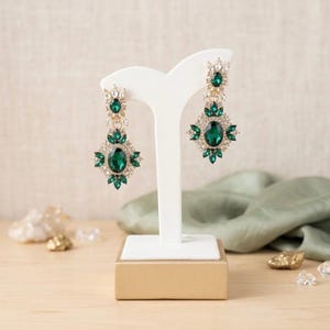 May include: A pair of dangling earrings featuring large oval-cut emerald green crystals surrounded by smaller clear rhinestones in a gold-toned metal setting. The earrings are displayed on a white T-shaped stand with a gold-colored rectangular base.