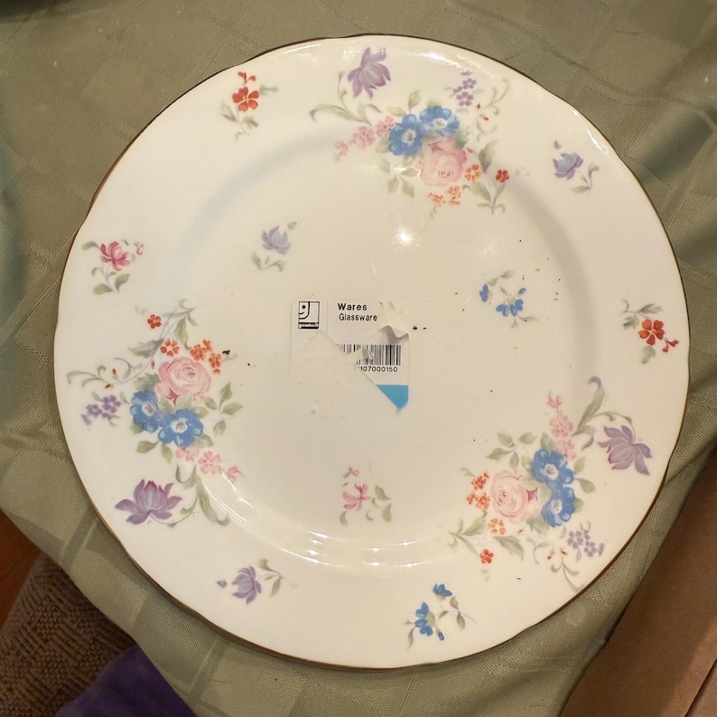Puede incluir: Un plato de cer&aacute;mica blanco con borde dorado, adornado con un estampado floral en tonos rosa, azul y morado. El plato tiene una etiqueta que dice "Wares Glassware".