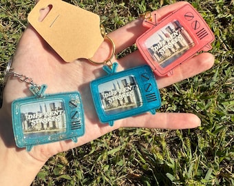 May include: Keychains in the shape of retro televisions in blue and pink. Each keychain has the words "Different Strokes" on the screen. The keychains are attached to a gold keyring and a brown tag.