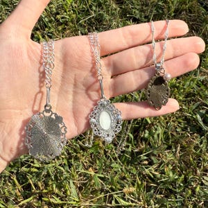 Puede incluir: Tres collares plateados con dise&ntilde;os de colgantes. El primer colgante tiene un dise&ntilde;o detallado en forma de sol. El segundo colgante tiene un centro ovalado blanco con un marco adornado. El tercer colgante tiene un dise&ntilde;o floral y una peque&ntilde;a perla.
