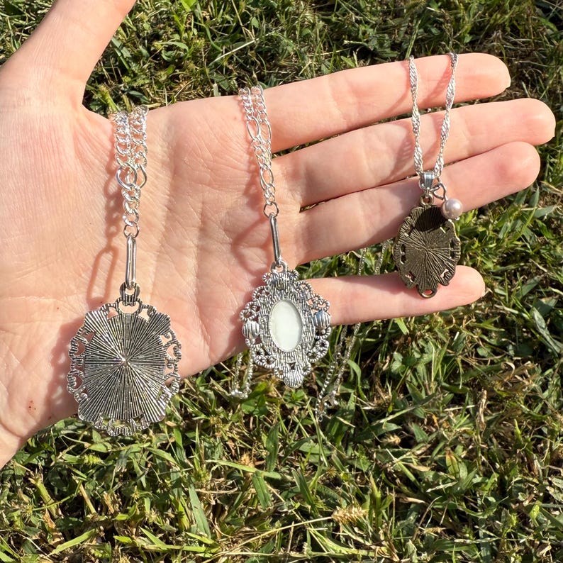 Puede incluir: Tres collares plateados con dise&ntilde;os de colgantes. El primer colgante tiene un dise&ntilde;o detallado en forma de sol. El segundo colgante tiene un centro ovalado blanco con un marco adornado. El tercer colgante tiene un dise&ntilde;o floral y una peque&ntilde;a perla.