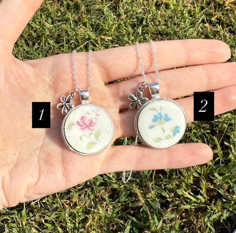 Puede incluir: Dos collares plateados con colgantes redondos con dise&ntilde;os florales. Un colgante tiene una flor rosa, el otro una flor azul. Cada collar tiene un peque&ntilde;o dije de flor y se muestra en una mano.