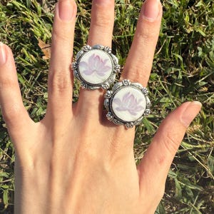 Puede incluir: Dos anillos plateados con un dise&ntilde;o floral. Cada anillo presenta una cara redonda y blanca con una flor de loto lavanda. Los anillos tienen un borde ornamentado. Los anillos est&aacute;n en una mano.