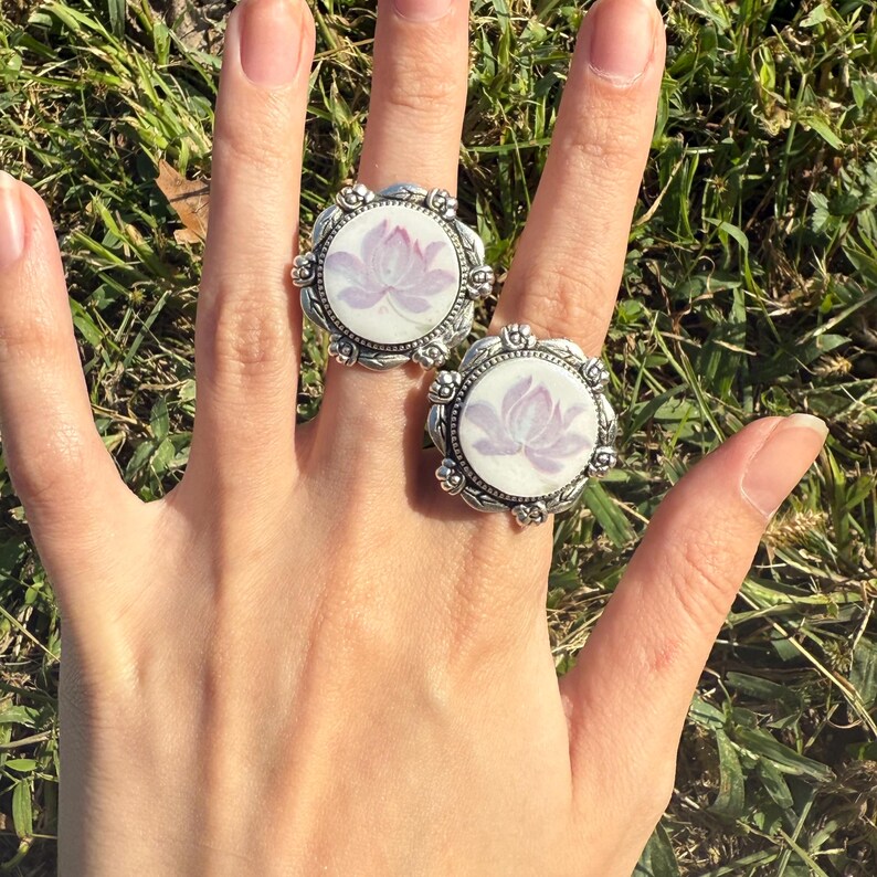Puede incluir: Dos anillos plateados con un dise&ntilde;o floral. Cada anillo presenta una cara redonda y blanca con una flor de loto lavanda. Los anillos tienen un borde ornamentado. Los anillos est&aacute;n en una mano.