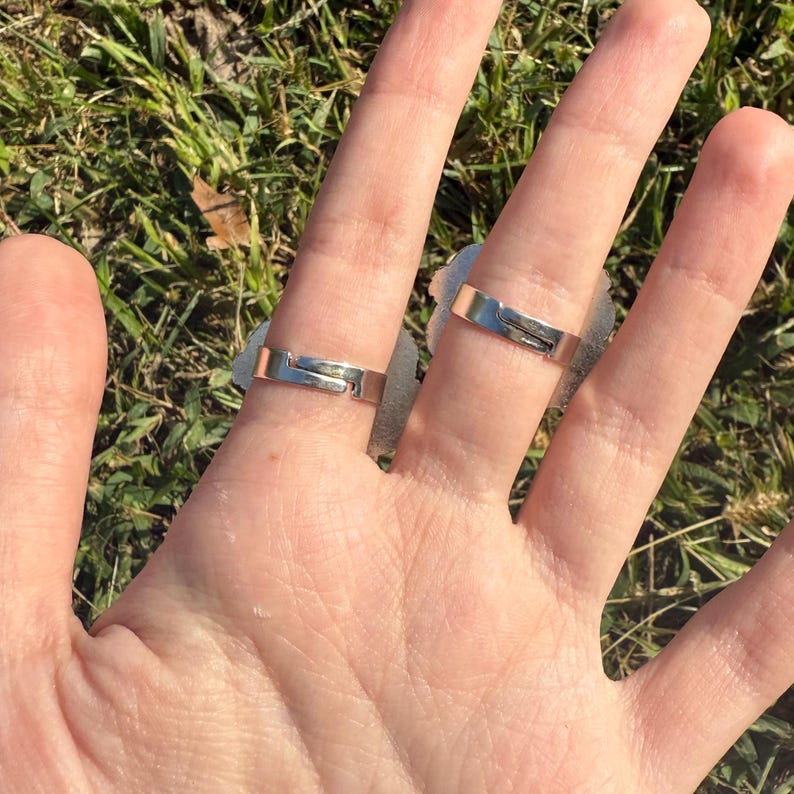 Puede incluir: Primer plano de una mano con dos anillos ajustables de plata. Los anillos tienen un dise&ntilde;o rectangular con un peque&ntilde;o espacio. El fondo es hierba verde borrosa.