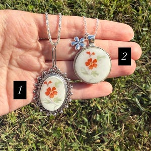 Puede incluir: Dos collares de plata con colgantes florales. Uno es ovalado con un marco adornado, el otro es redondo con un dije de flor. Ambos colgantes presentan flores naranjas sobre un fondo blanco, con cadenas de plata.