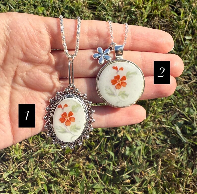 Puede incluir: Dos collares de plata con colgantes florales. Uno es ovalado con un marco adornado, el otro es redondo con un dije de flor. Ambos colgantes presentan flores naranjas sobre un fondo blanco, con cadenas de plata.