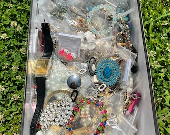 Puede incluir: Una caja de plástico transparente llena de una variedad de joyas, incluyendo collares, pulseras, aretes y un reloj. Las joyas están dispuestas en un patrón aleatorio e incluyen una variedad de colores y estilos.