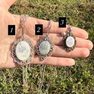Puede incluir: Tres collares con colgantes ovalados con dise&ntilde;os florales. Los colgantes tienen un fondo blanco con flores moradas. Los dos primeros colgantes tienen un marco de filigrana plateada, mientras que el tercero tiene un marco de bronce y una peque&ntilde;a perla. Los collares se exhiben sobre un fondo de hierba.