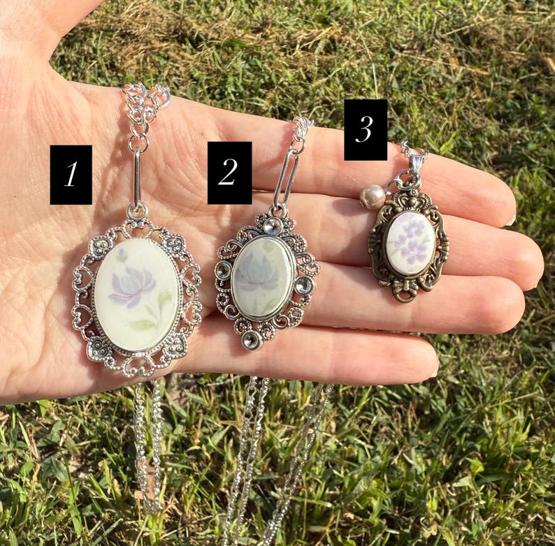 Puede incluir: Tres collares con colgantes ovalados con dise&ntilde;os florales. Los colgantes tienen un fondo blanco con flores moradas. Los dos primeros colgantes tienen un marco de filigrana plateada, mientras que el tercero tiene un marco de bronce y una peque&ntilde;a perla. Los collares se exhiben sobre un fondo de hierba.