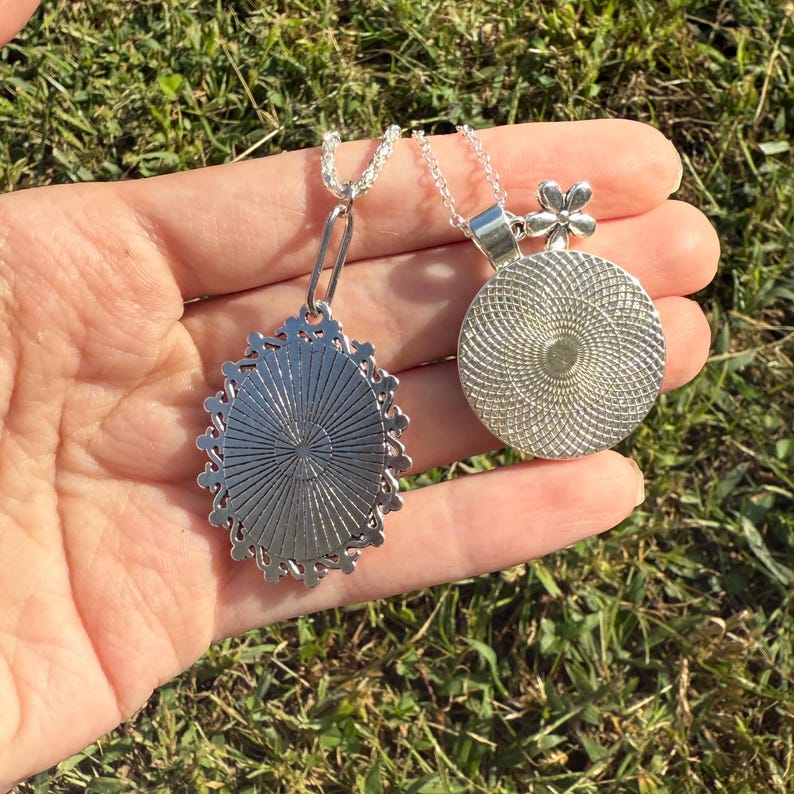 Puede incluir: Dos collares plateados con colgantes. Uno es ovalado con borde decorativo y patr&oacute;n radial. El otro es redondo con un adorno floral y dise&ntilde;o en espiral. Ambos collares se muestran sobre un fondo de hierba verde.