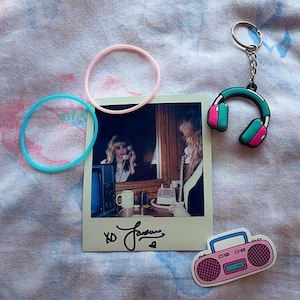 Puede incluir: Un surtido de artículos de temática retro. Una foto Polaroid con una mujer al teléfono, dos pulseras de goma, un llavero con auriculares y una pegatina de radio rosa. La foto tiene una firma y el texto "XO".