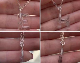 Puede incluir: Un collar de cadena plateada con un colgante transparente que presenta la letra "E" en su interior. El colgante está lleno de texto impreso pequeño y negro.