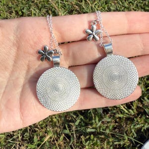 Puede incluir: Dos collares colgantes plateados con un dije floral y un colgante circular con un dise&ntilde;o en espiral. Los collares se muestran en una mano sobre un fondo de hierba verde.