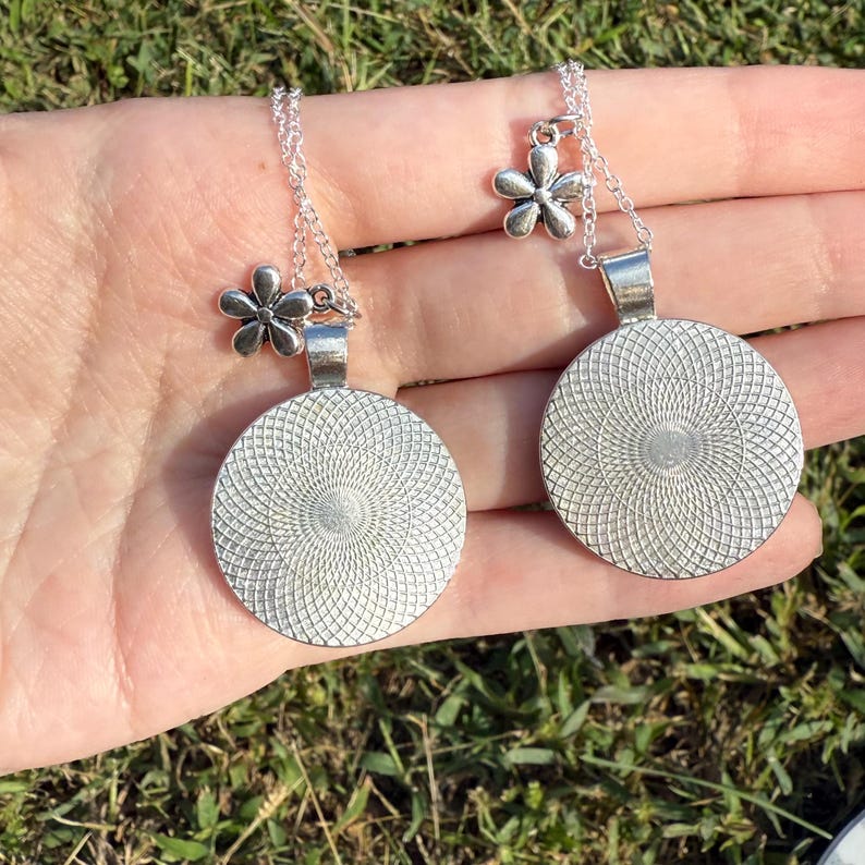 Puede incluir: Dos collares colgantes plateados con un dije floral y un colgante circular con un dise&ntilde;o en espiral. Los collares se muestran en una mano sobre un fondo de hierba verde.