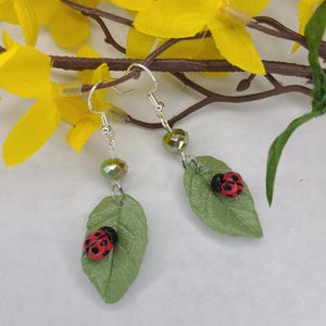 May include: A pair of dangle earrings featuring silver-toned hooks, a faceted olive-green bead, and a textured light green leaf charm. Each leaf is adorned with a small red ladybug with black spots. The earrings hang from thin silver-toned wires.