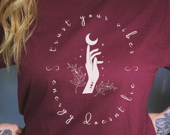 May include: Burgundy t-shirt with a white graphic design featuring a hand holding a crescent moon and stars. The text around the graphic reads "Trust your vibes, energy doesn't lie."
