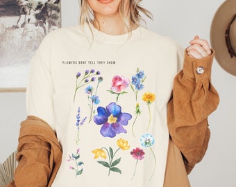 Puede incluir: Una camiseta de color crema con un diseño floral de acuarela y el texto "Flowers don't tell, they show".