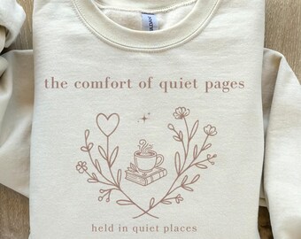 Può includere: Felpa color crema con la scritta "the comfort of quiet pages" e un disegno floreale con un cuore, una tazza di caffè e un libro. Le maniche sono lunghe e il materiale sembra morbido.