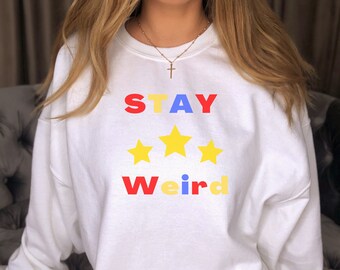 May include: White sweatshirt with the text "STAY Weird" in colorful letters and three yellow stars.
