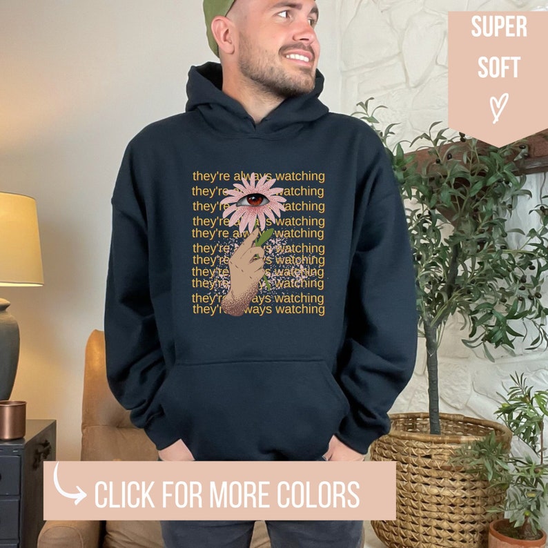 May include: Black hooded sweatshirt with a pink flower graphic and the text "they're always watching" repeated multiple times. The flower has an eye in the center.