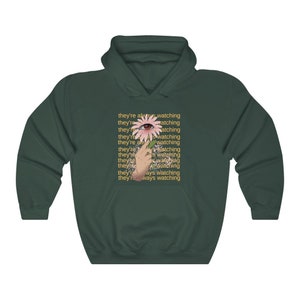 May include: A dark green hoodie with a pink flower and an eye in the center. The text "they're always watching" is repeated multiple times in yellow.