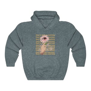 May include: A dark gray hoodie with a pink flower with an eye in the center. The text "they're always watching" is repeated multiple times in yellow.
