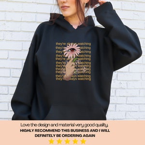 May include: Black hooded sweatshirt with a pink flower with an eye in the center. The text "they're always watching" is repeated in a pattern on the sweatshirt.