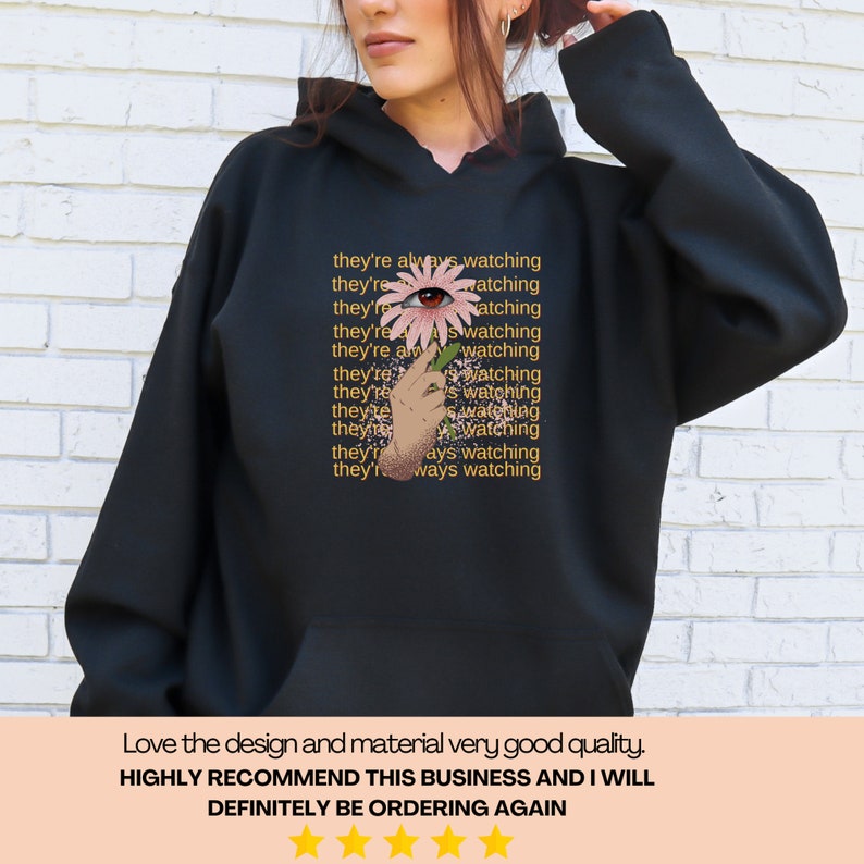 May include: Black hooded sweatshirt with a pink flower with an eye in the center. The text "they're always watching" is repeated in a pattern on the sweatshirt.