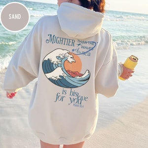 Könnte beinhalten: Ein beiger Hoodie mit einem Grafikdruck einer blauen Welle und einer untergehenden Sonne. Der Text "MIGHTER THAN THE WAVES OF THE SEA is his love for you Psalm 93:4" ist auf der Rückseite aufgedruckt.