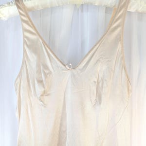 May include: A vintage, cream-colored slip dress with a V-neckline and spaghetti straps. The dress features a delicate bow detail at the bust and a flowing, sheer skirt. The slip is hanging on a white padded hanger.