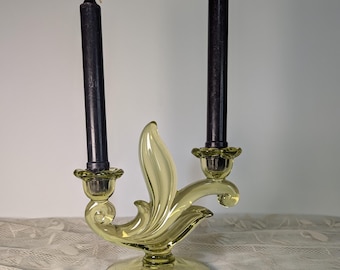 May include: A pale yellow glass candelabra with two black candles. The candelabra has a decorative, swirling design and a round base. The candles are tall and slender, with a dark, matte finish. The overall style is vintage.
