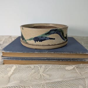 May include: A handmade ceramic bowl with a beige exterior and a dark brown interior. The bowl features a blue and green abstract design around the exterior. The bowl is sitting on top of two blue books.