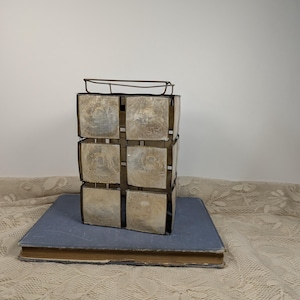 May include: A decorative, vintage metal and shell cube-shaped object with a rectangular top, resting on a blue book. The cube is composed of six square shell panels, framed by a dark metal. The book has a blue cover and aged, yellowed pages.
