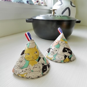 May include: Two cone-shaped pot holders with a cat pattern in yellow, black, and white. Each pot holder has a loop with red, white, and blue stripes. A black cast iron pot sits in the background.