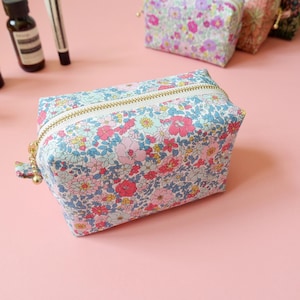 May include: A floral print cosmetic bag with a gold zipper. The bag features a pattern of pink, blue, and yellow flowers on a white background. Other similar bags are visible in the background.