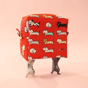 Op de afbeelding: Rode stoffen toilettas met een herhalend patroon van katten in verschillende poses. De tas staat op twee grijze kattenfiguren.