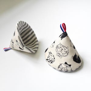 以下が含まれることがあります： 猫のイラストが描かれたベージュ色の鍋つかみ2つ。1つは白黒のストライプ柄、もう1つは様々な猫の顔が描かれています。どちらも、吊り下げ用の赤、白、青のトリムが付いた小さなループが付いています。