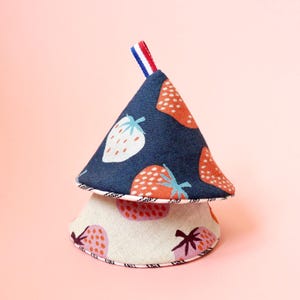 May include: Two fabric pot holders, one atop the other, with a strawberry theme. The top pot holder is navy blue with large red and white strawberries. The bottom pot holder is off-white with pink and purple strawberries. A red, white, and blue ribbon is attached to the top.