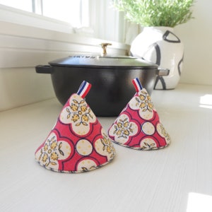 May include: Two red and white pot holders with a floral pattern. The pot holders are shaped like cones and have a blue and white ribbon tied around the top.