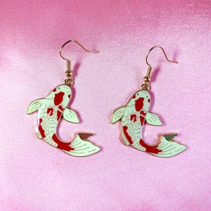 May include: A pair of gold-tone earrings featuring a white and red koi fish design. The fish are facing opposite directions and have a detailed, painted finish.