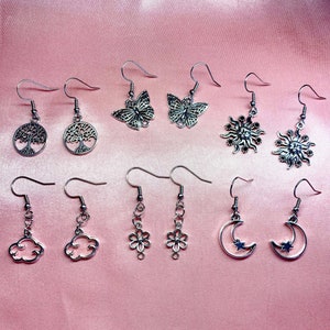 Puede incluir: Un conjunto de 10 pendientes de plata con varios diseños, incluyendo árboles, mariposas, soles, nubes, flores y lunas. Los pendientes son todos de plata y tienen un aspecto delicado y femenino.