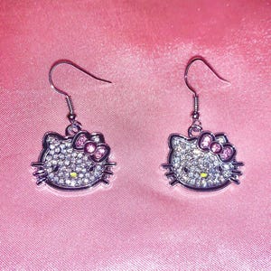 Pode incluir: Um par de brincos Hello Kitty prateados com laços rosa e strass transparentes.