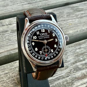 Puede incluir: Un reloj de pulsera Oris vintage con caja redonda plateada y esfera negra. La esfera presenta números arábigos blancos, un calendario perimetral con una aguja de punta roja y una subesfera. Incluye una correa de cuero de color marrón.