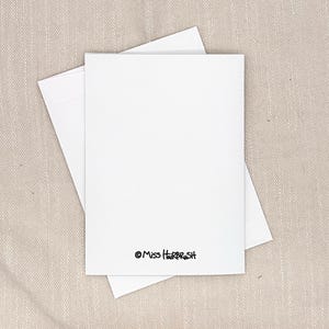 以下が含まれることがあります： 無地の白い長方形のグリーティングカードと、その下に置かれたお揃いの白い封筒。カードの下部中央には、著作権記号を伴う黒い文字で「Miss HairBrush」と印刷されています。