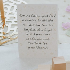 May include: A cream-colored card with text in elegant script, encouraging the creation of a baby keepsake. The card features a subtle pattern of baby-themed illustrations. Wooden blocks are visible in the foreground.