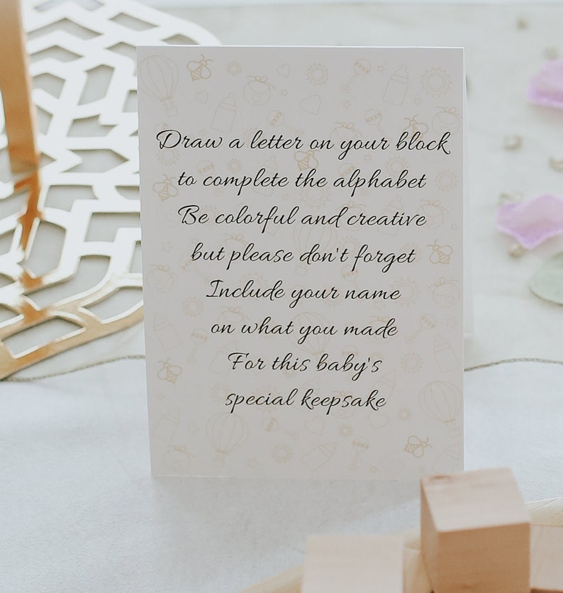 May include: A cream-colored card with text in elegant script, encouraging the creation of a baby keepsake. The card features a subtle pattern of baby-themed illustrations. Wooden blocks are visible in the foreground.