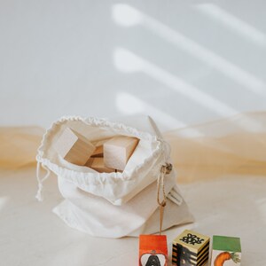 May include: Wooden alphabet blocks with hand-painted letters and illustrations, including an apple, a bee, and a carrot. The blocks are displayed with a drawstring bag filled with plain wooden blocks, perfect for educational play.