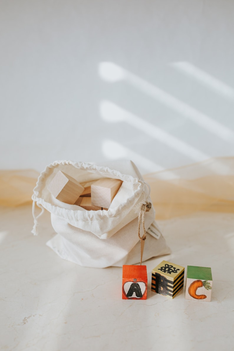 May include: Wooden alphabet blocks with hand-painted letters and illustrations, including an apple, a bee, and a carrot. The blocks are displayed with a drawstring bag filled with plain wooden blocks, perfect for educational play.